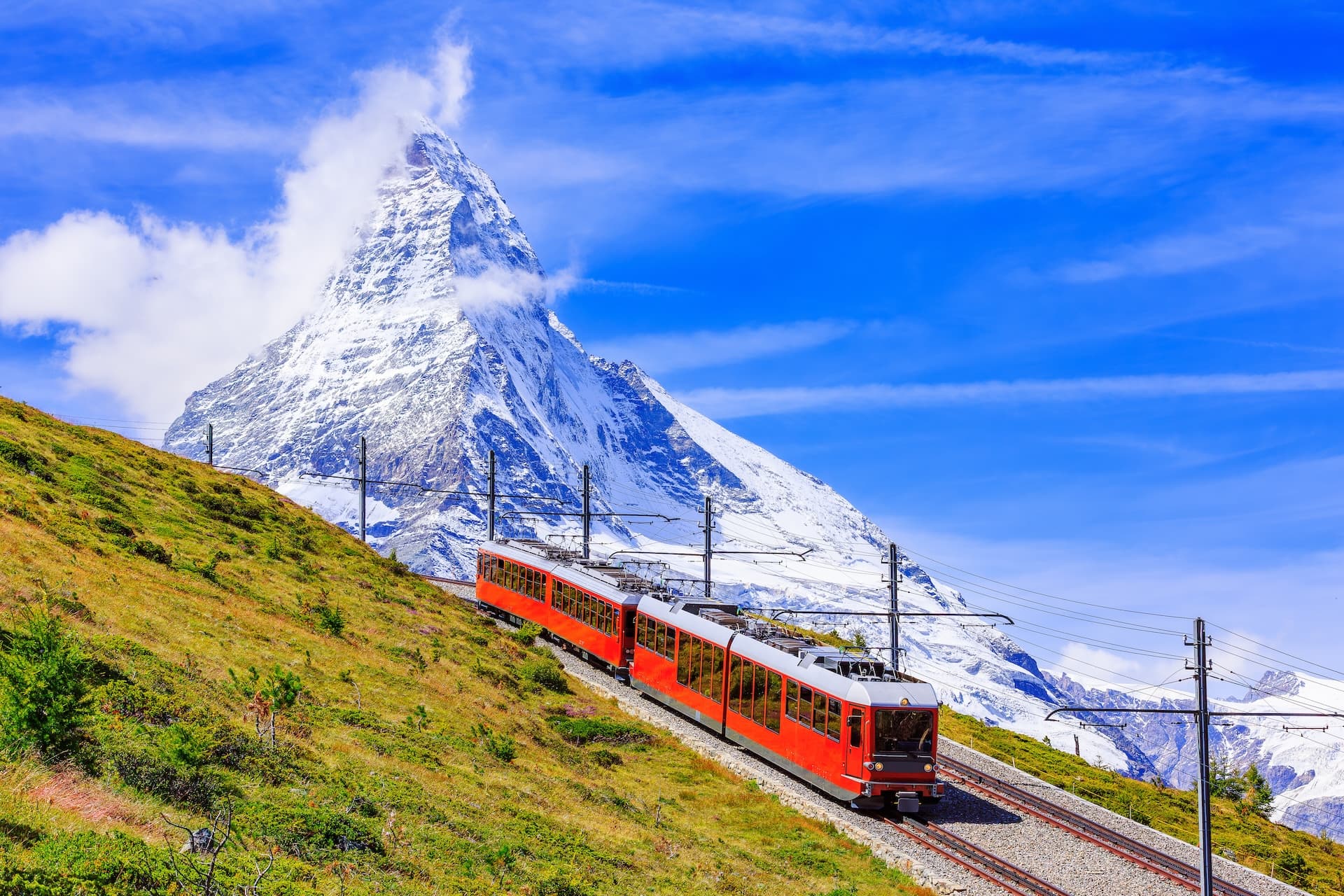 Red Gornergrat cogwheel train ascending grassy slope below snow-capped Matterhorn peak.
