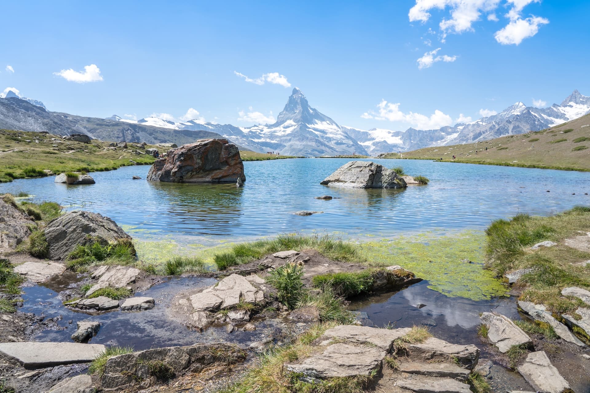 Alpine lake with Matterhorn peak in distance, surrounded by rocky shore and grassy hills.