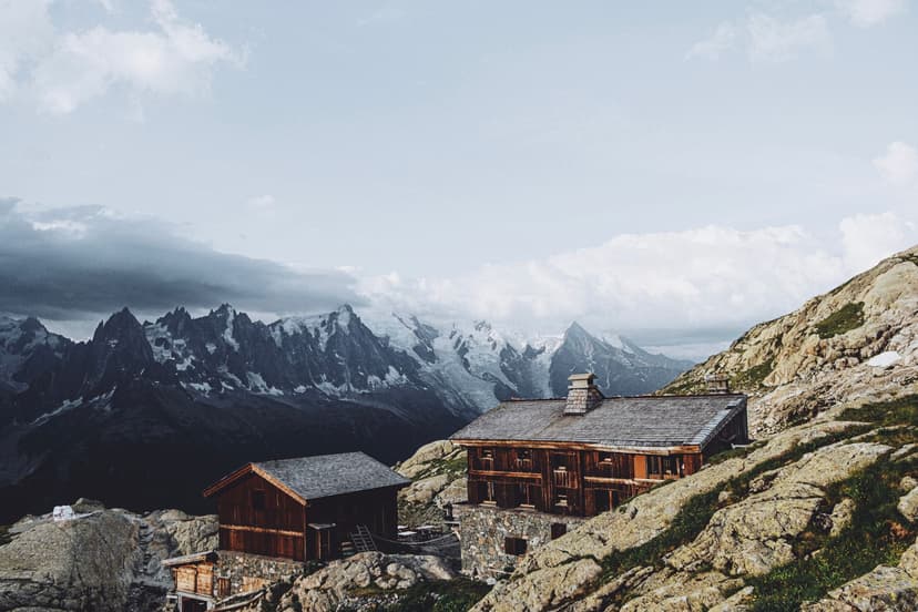 Wooden refuge buildings on rocky terrain with snowy alpine peaks under cloudy sky, Refuge de Lac Blanc.