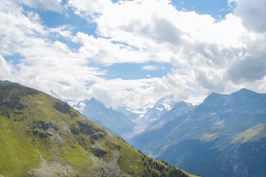 Alpine mountain range with grassy foreground and snow-capped peaks under cloudy blue sky, Zinal to Weisshorn.