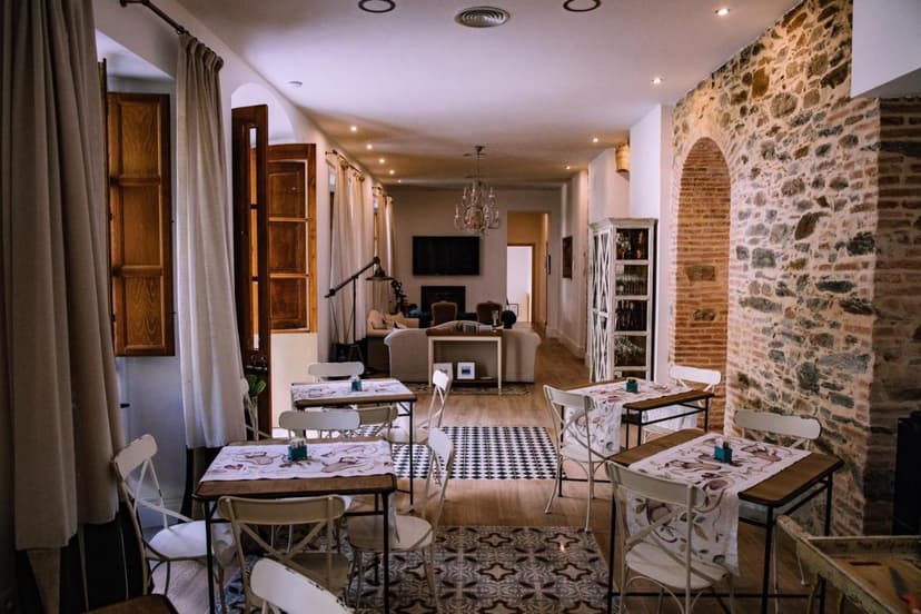 Dining room at Hotel Luz Almonaster with stone wall, white chairs, and view into a lounge area.