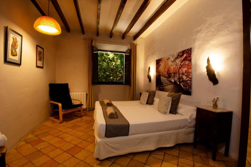 Twin beds in rustic room with terracotta floor, exposed beams, and window view of greenery at S´Hostal d´Esporles.