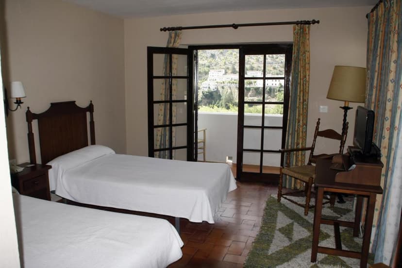 Twin bedroom with balcony view of white buildings on a green hillside, Villa Turística de Grazalema.