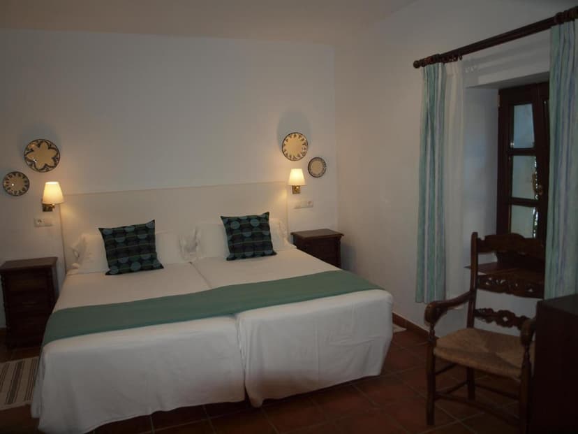 Twin beds with white linens and teal accents in a room with terracotta floors and a wooden chair.
