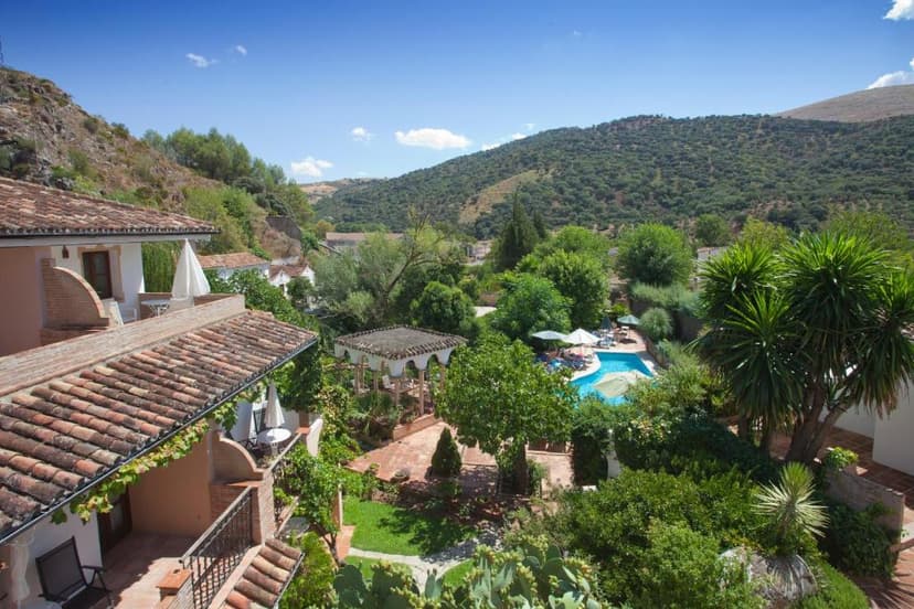 Terracotta roof buildings overlooking a lush garden and swimming pool with wooded hills under blue sky.