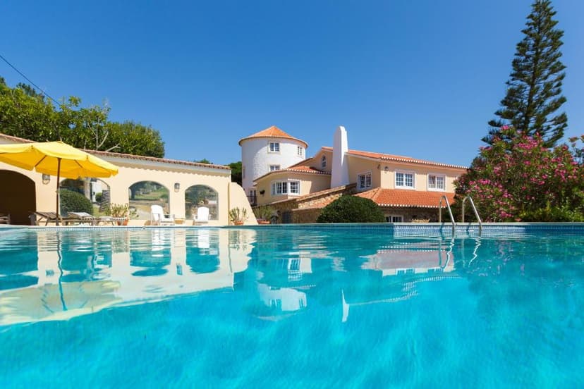 Swimming pool reflecting guesthouse with yellow umbrella and flowering tree under blue sky.