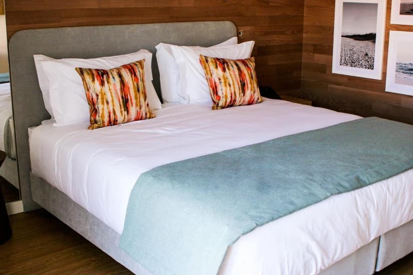 Hotel bedroom with white linens, patterned pillows, and beach artwork on wood paneling in Cascais.