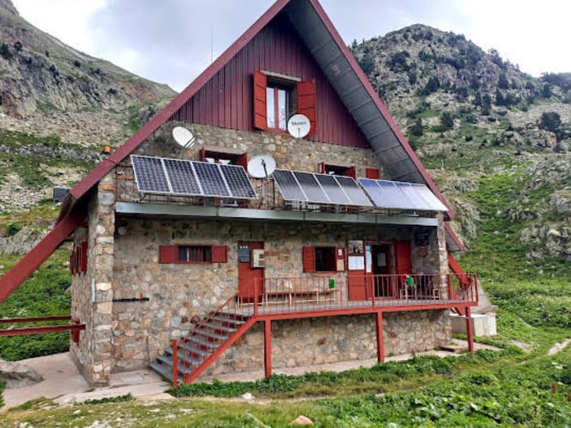 Stone mountain refuge Refugio de Respomuso with solar panels and red trim against green slopes.