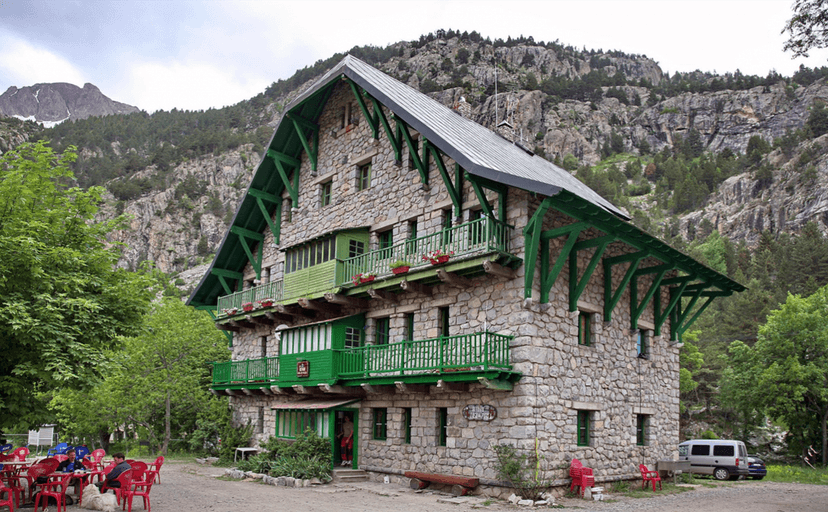 Stone mountain hut with green trim, outdoor seating, and forested rocky peaks.