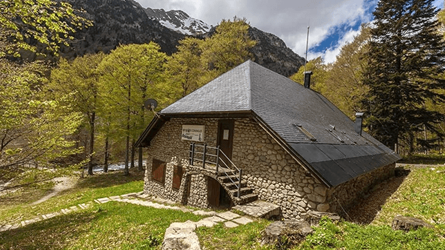 Stone refuge Refugio Conangles surrounded by green trees with snow-capped mountains in the background