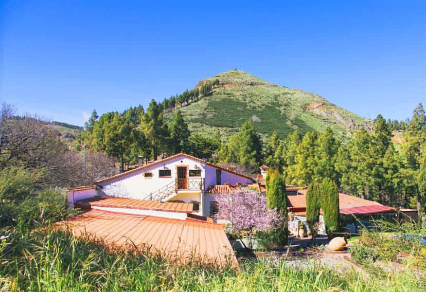 Rural hotel building with terracotta roof set against a steep, green mountain under a clear blue sky.