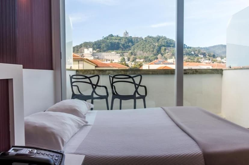Hotel room with bed and balcony view of green hill and town rooftops, possibly Viana.