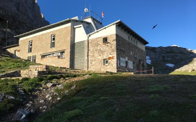 Stone mountain refuge Vega de Urriellu with grassy slope and rocky peaks under blue sky.