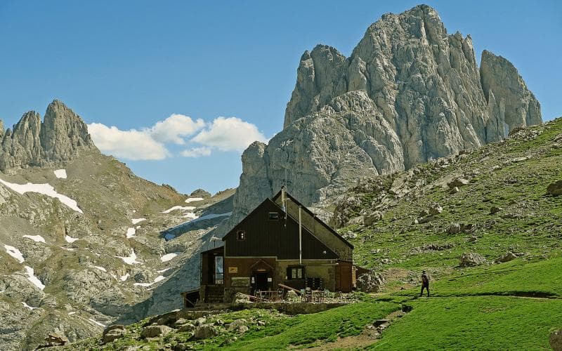 Mountain refuge hut below jagged peaks with snow patches and a hiker on green slope.