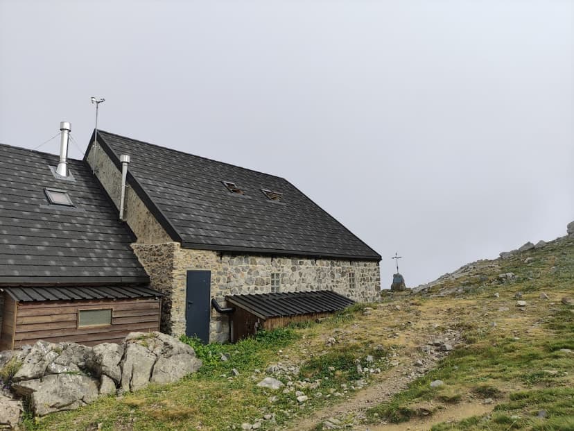 Stone mountain refuge with dark roof next to rocky path under overcast sky, Refugio Collado Jermoso