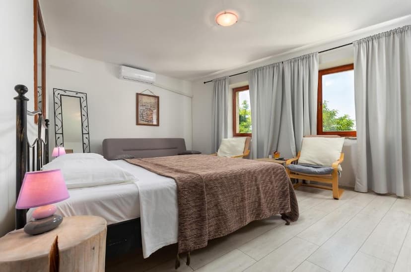 Bedroom with white walls, wood floor, brown blanket, and windows showing greenery.