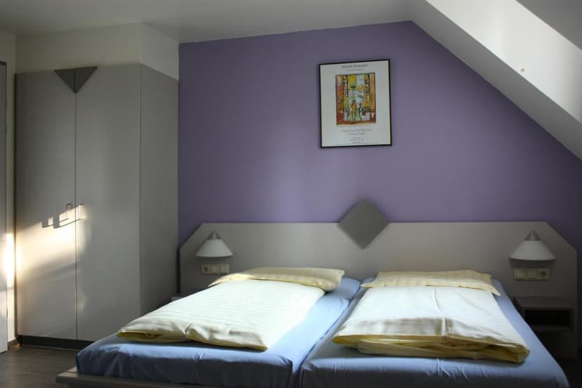 Twin beds in hotel room with lavender wall, white sloped ceiling, and light gray wardrobe.