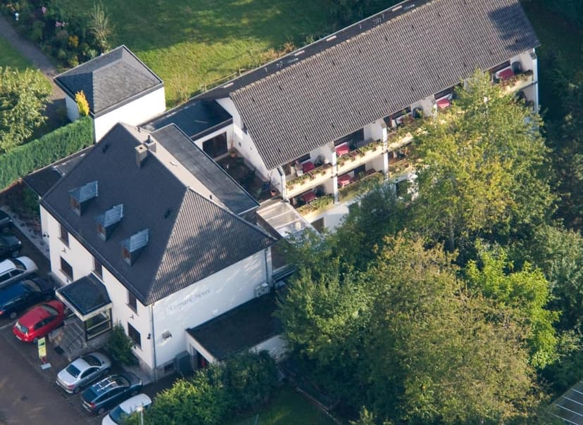 Aerial view of Kurparkhotel Gemünd building with dark roof, surrounded by green trees and parked cars.