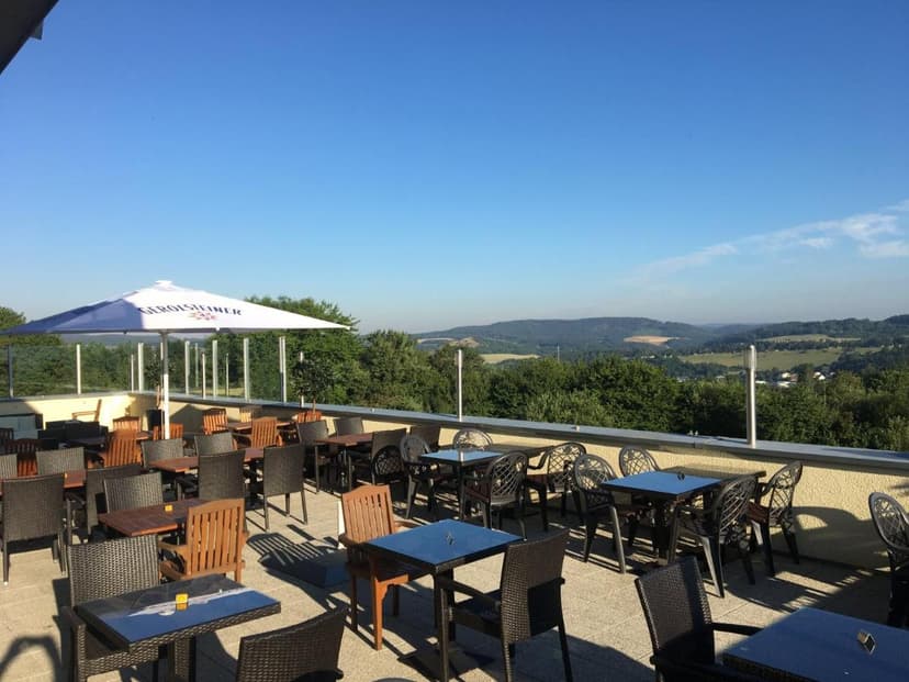 Outdoor terrace dining area with tables and chairs overlooking rolling green hills under a clear blue sky.