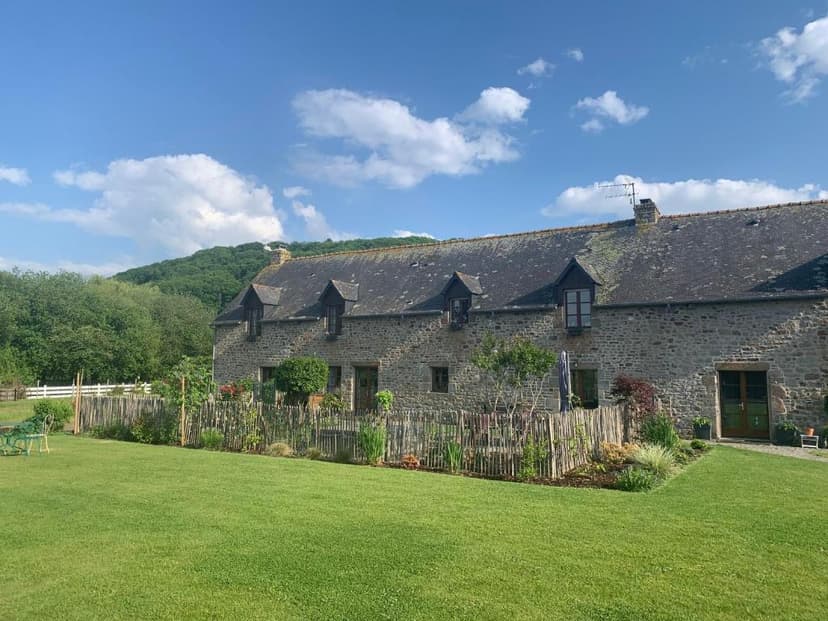 Stone gîte with dormer windows and garden, set against a wooded hillside under blue sky.