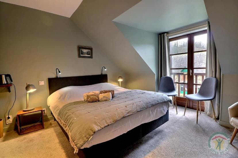 Bedroom with sloped ceiling, large bed, and double window doors with view of greenery.