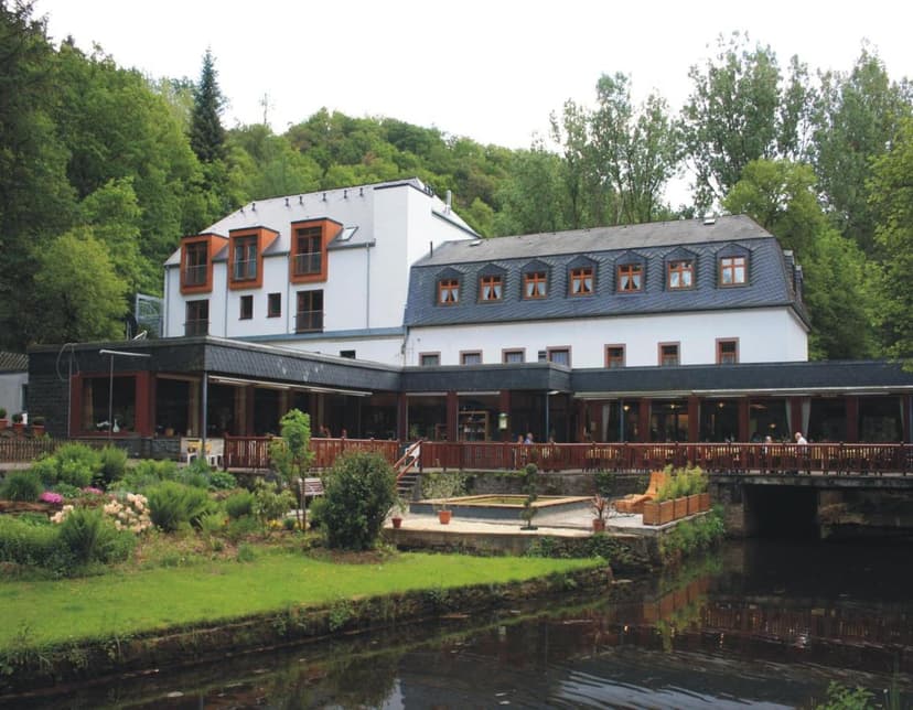 White building with dark roof next to a river, surrounded by lush green forest.