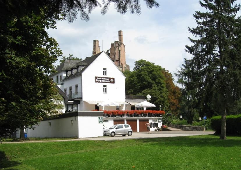 Hotel-Restaurant Burg Ramstein with ruins visible behind lush trees and green lawn.