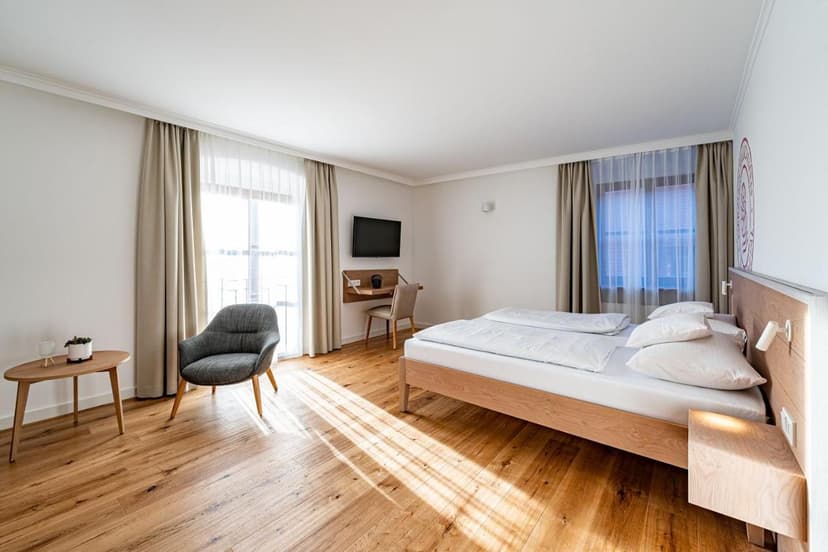 Bright hotel room with wooden floor, double bed, gray armchair, and wall-mounted TV at Schattenhofer Braugasthof.