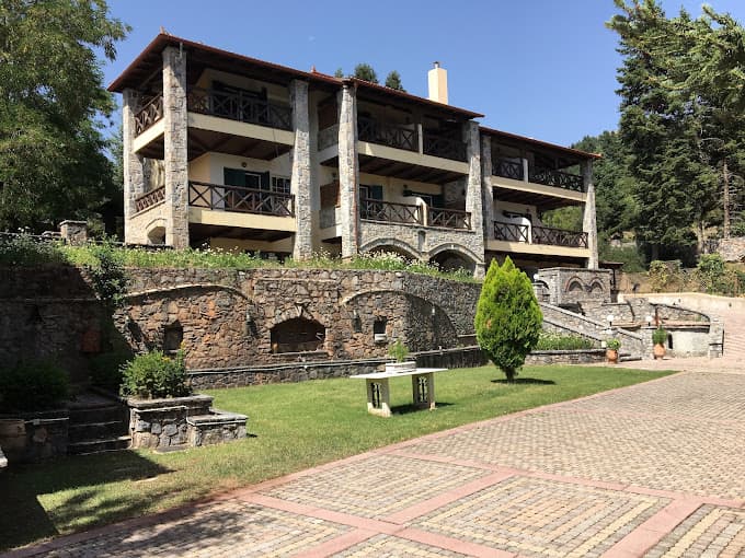 Stone chalet with balconies above a grassy area and paved courtyard, surrounded by trees.
