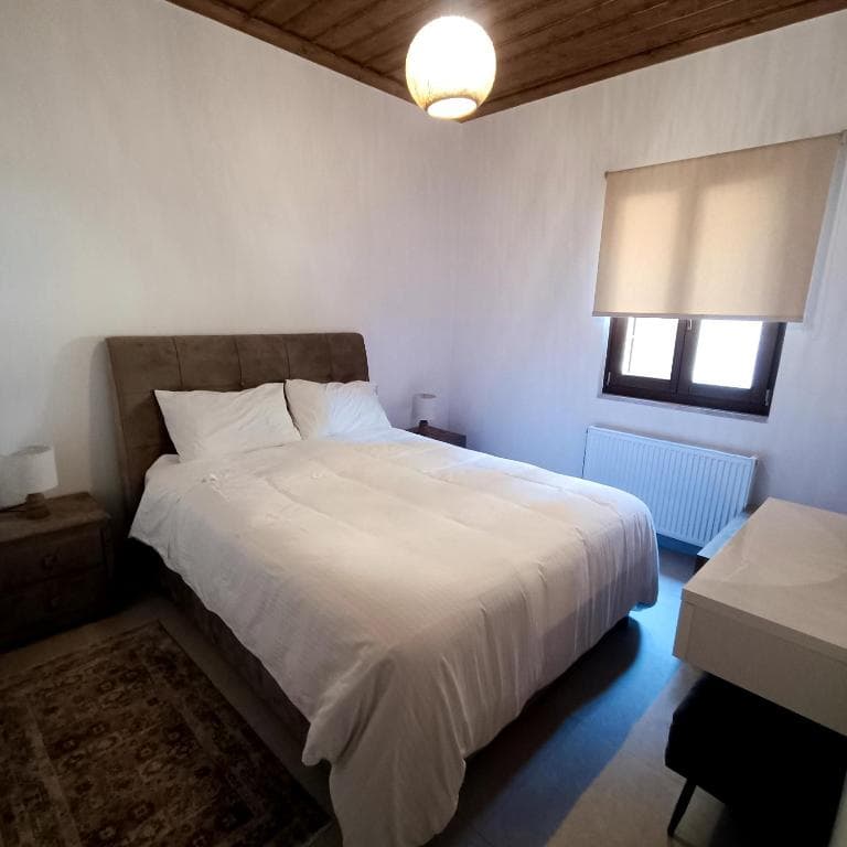 Bedroom with large bed, white linens, wooden ceiling, and window with roller blind.