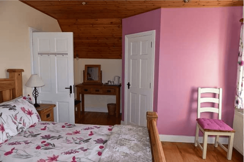 Bedroom with wooden ceiling, pink wall, floral bedding, and white wooden chair.
