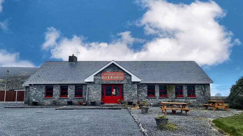 Cappabhaile House Bed & Breakfast with stone facade, red door, and picnic tables under blue sky.