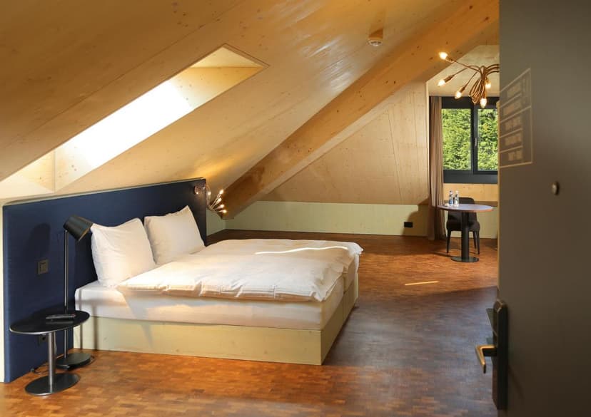 Attic hotel room with sloped wood ceilings, white bedding, and view of green trees.