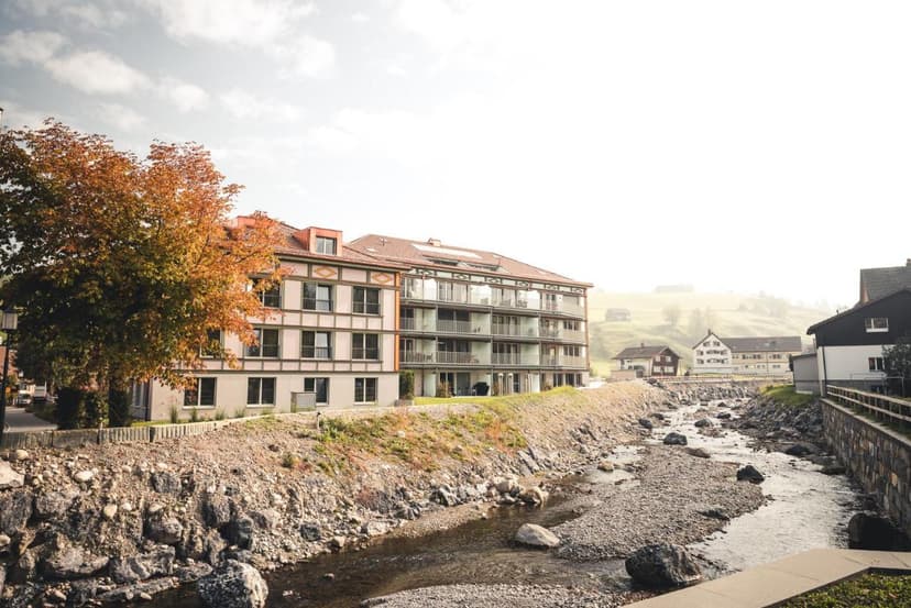 Modern lodge building next to a rocky stream with autumn foliage in a misty alpine village setting.