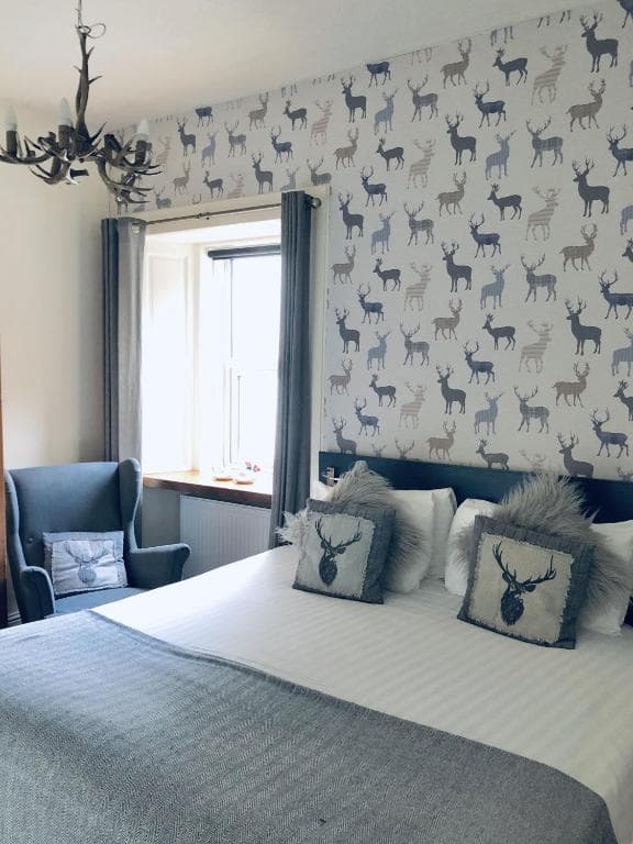 Bedroom with stag-pattern wallpaper, antler chandelier, and grey armchair near a window.