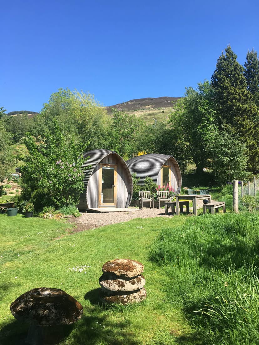 Two wooden glamping pods on green grass with hills and blue sky, near Loch Tay.