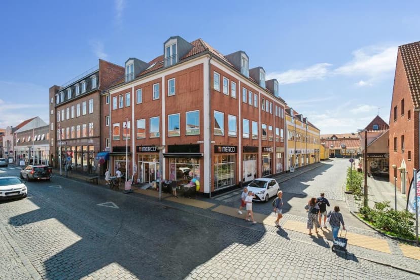 Cobblestone street in Rønne with brick buildings, Imerco store, and pedestrians on a sunny day.
