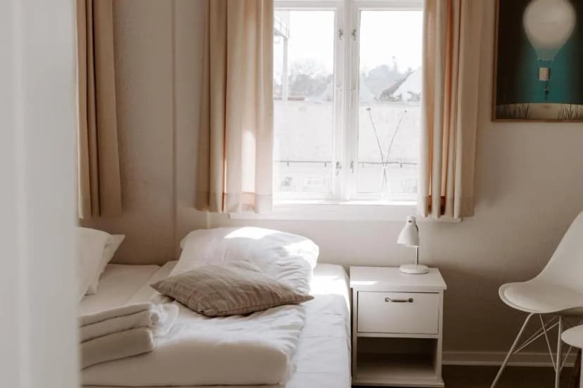 Brightly lit hotel room with white bedding, nightstand, and window view in Sandvig Havn.