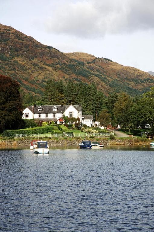 White hotel by lake with small boats, backed by large brown and green mountains