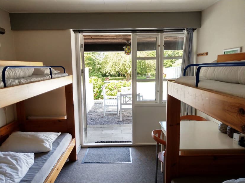 Hostel room with wooden bunk beds opening to a sunny garden patio with white chairs