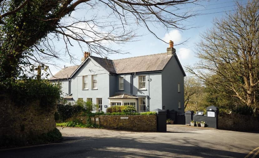 Light blue country house with stone wall entrance framed by large trees under a bright sky.