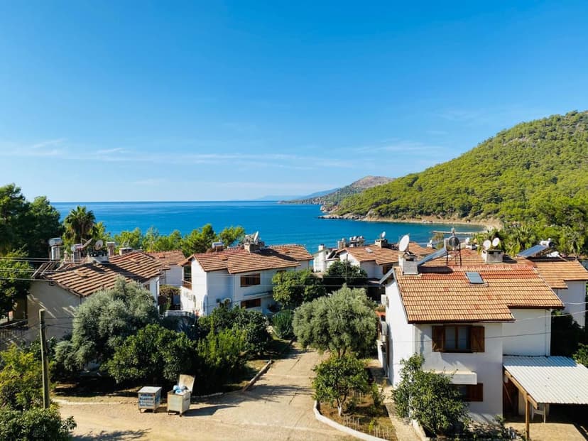 White houses with terracotta roofs overlooking a bright blue sea and green forested hills.