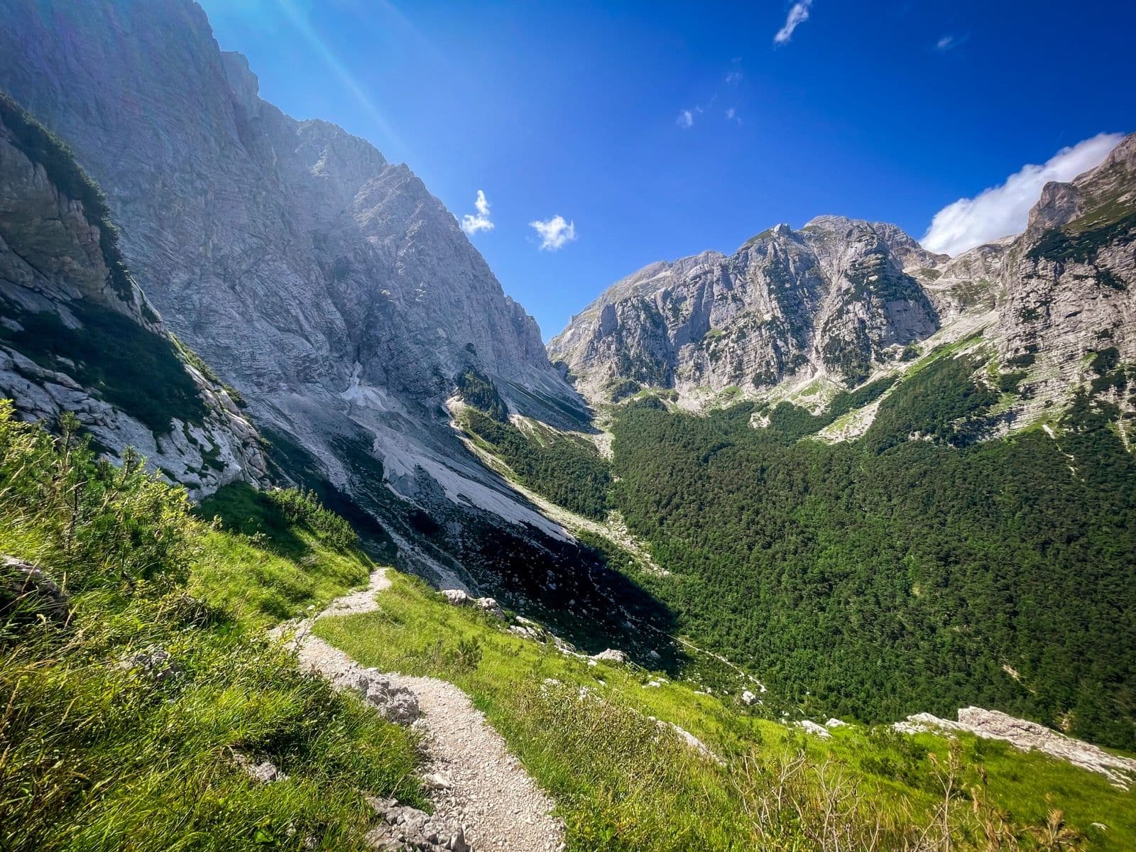 Hiking trail winding through grassy slope between steep gray mountains under blue sky.