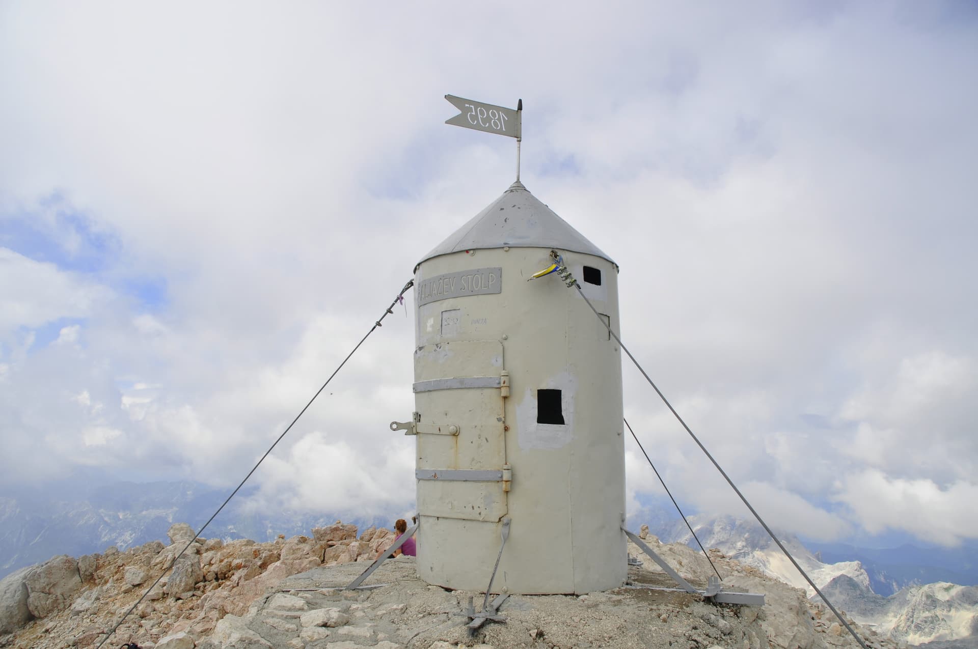 Aljažev Stolp summit shelter on rocky mountain peak with cloudy sky and distant peaks