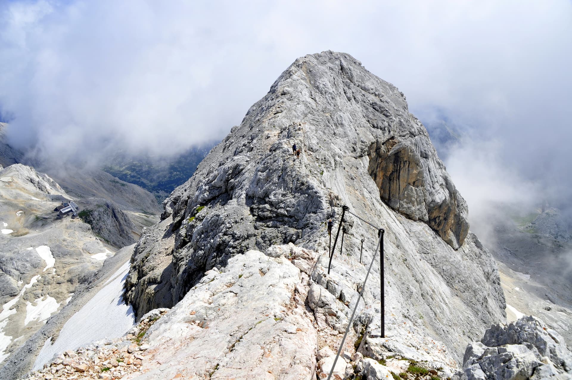 Hikers ascending steep, rocky Triglav mountain trail with fixed cables, partially obscured by clouds.