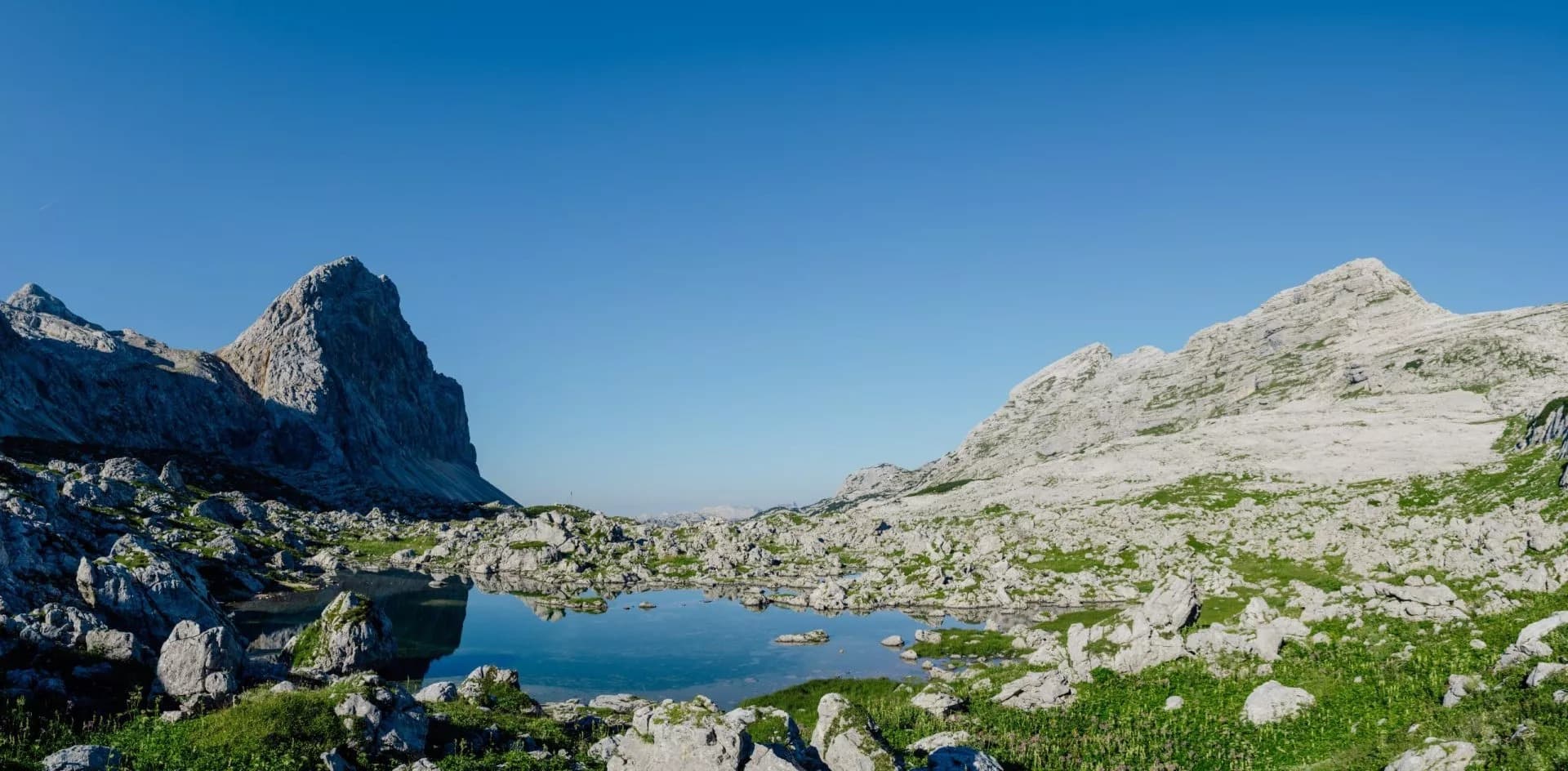 Alpine lake reflecting jagged mountains surrounded by rocks and green grass under a clear blue sky.