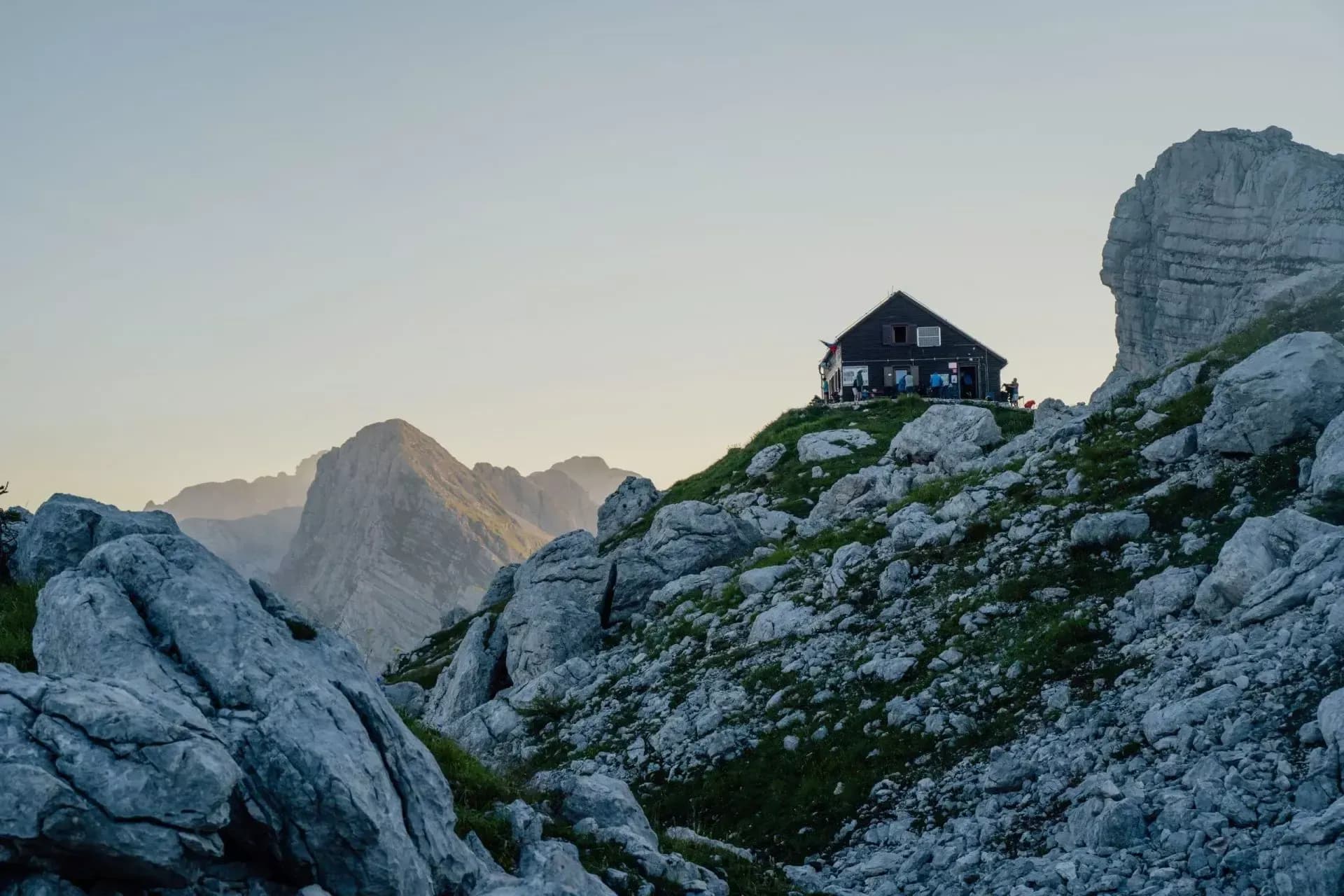 Mountain hut on rocky slope with hikers, set against distant peaks at dusk.
