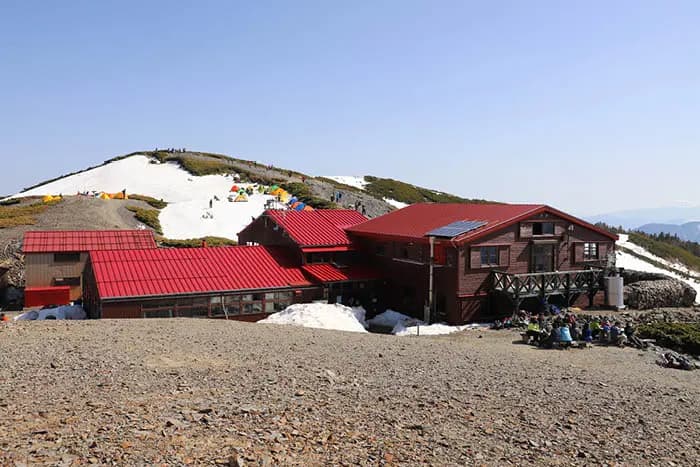 Chogatake mountain hut with red roof near snow patches and camping tents on a sunny day.