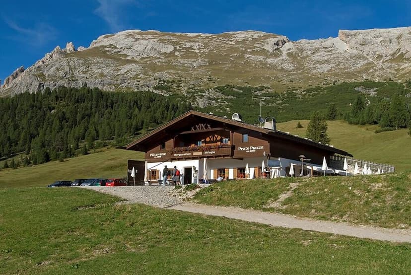 Rifugio Prato Piazza mountain hut with outdoor seating below rocky peaks and green slopes.