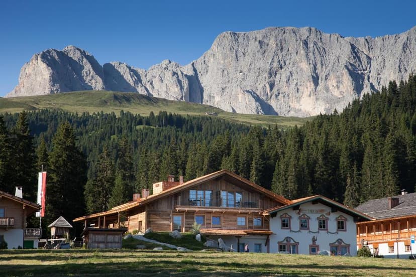 Alpine hotel complex with wooden chalets nestled below massive gray mountains and dense pine forest.